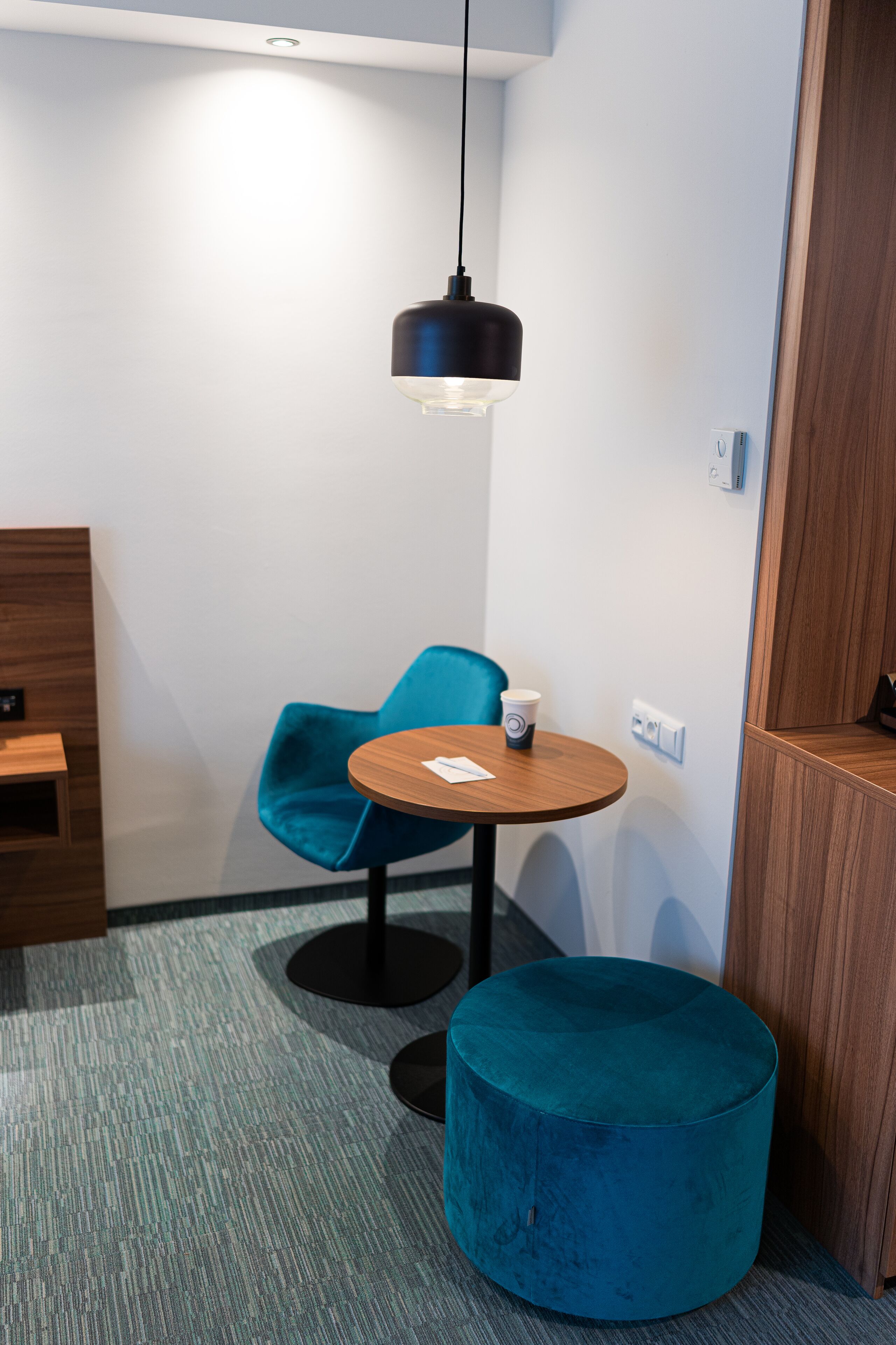 Una habitación pequeña y moderna con una mesa redonda de madera, una silla color esmeralda y un reposapiés a juego. Una lámpara colgante negra ilumina la mesa.