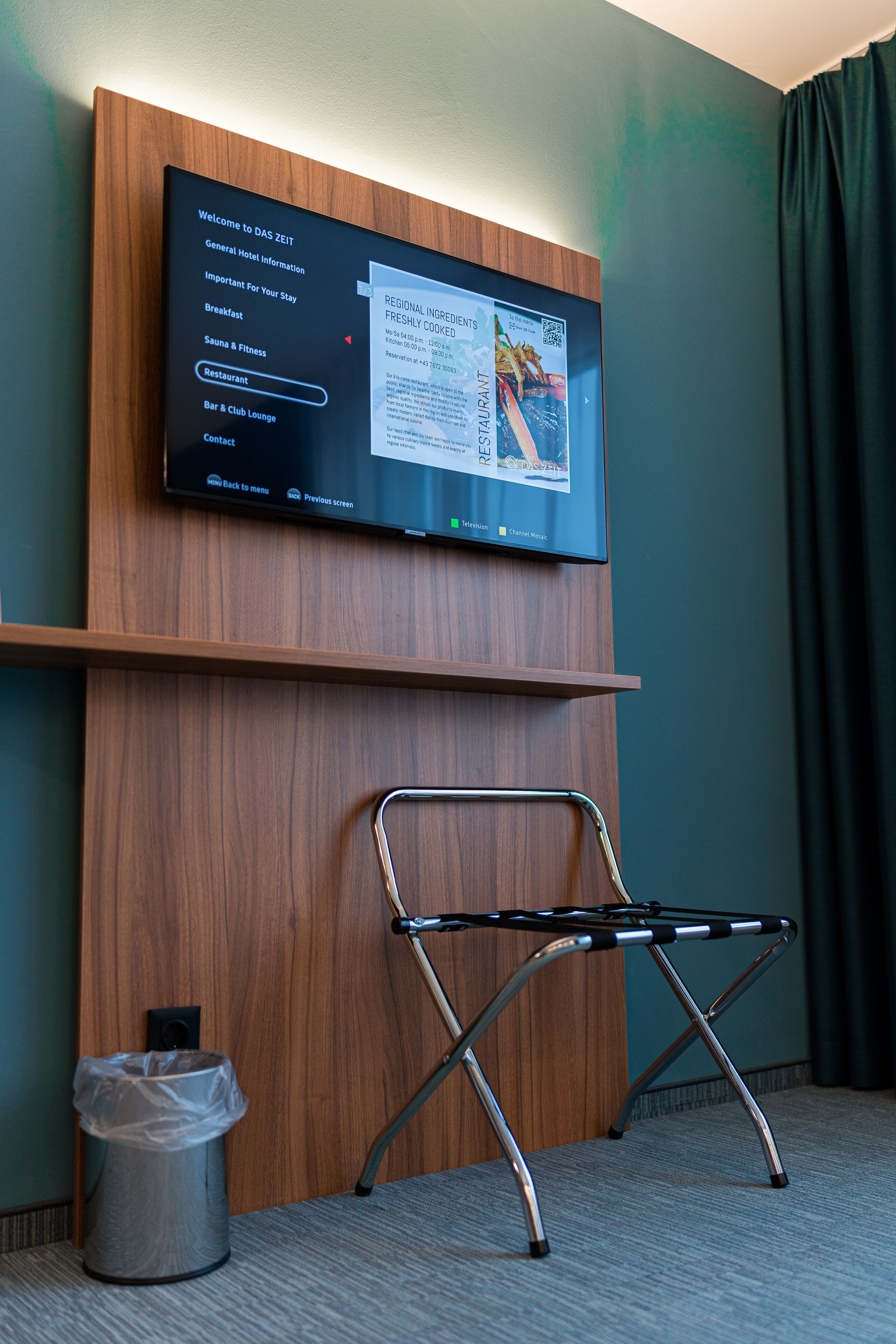 A hotel room with a mounted TV displaying a menu, a foldable chair, a trash bin, and a wooden panel wall.