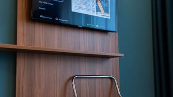 A hotel room with a mounted TV displaying a menu, a foldable chair, a trash bin, and a wooden panel wall.
