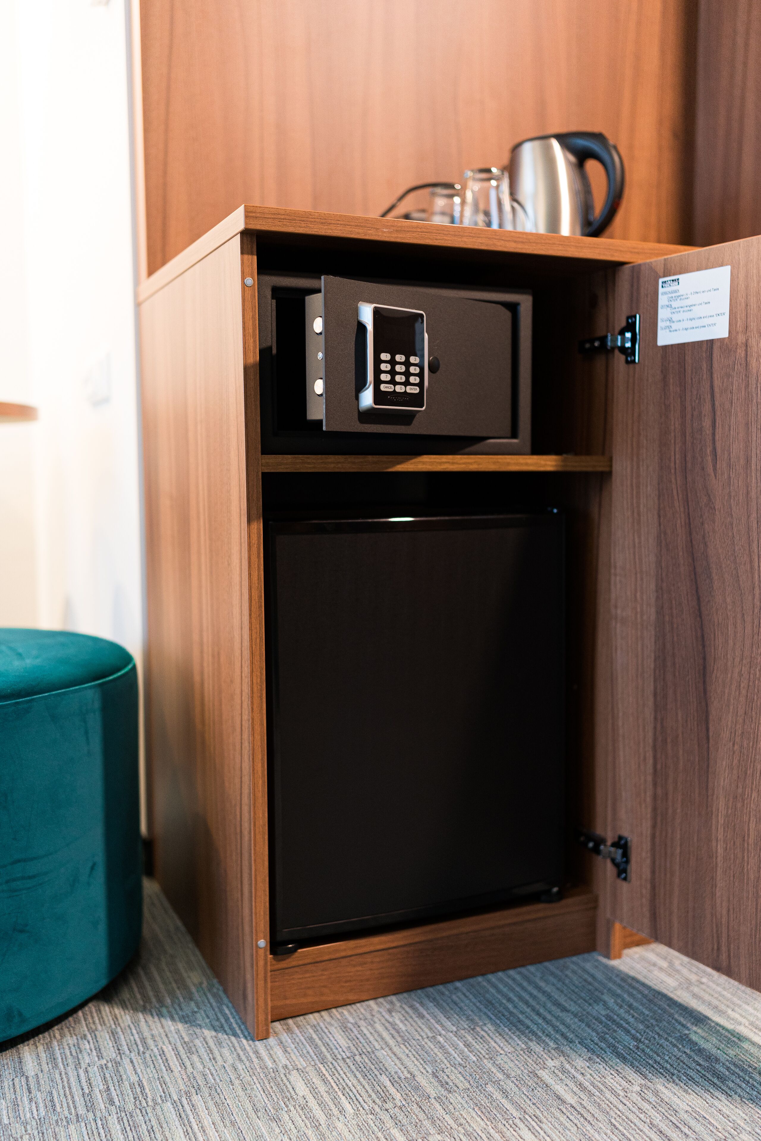 A compact minibar with a microwave and a small refrigerator, a teal ottoman, and a wooden panel wall.