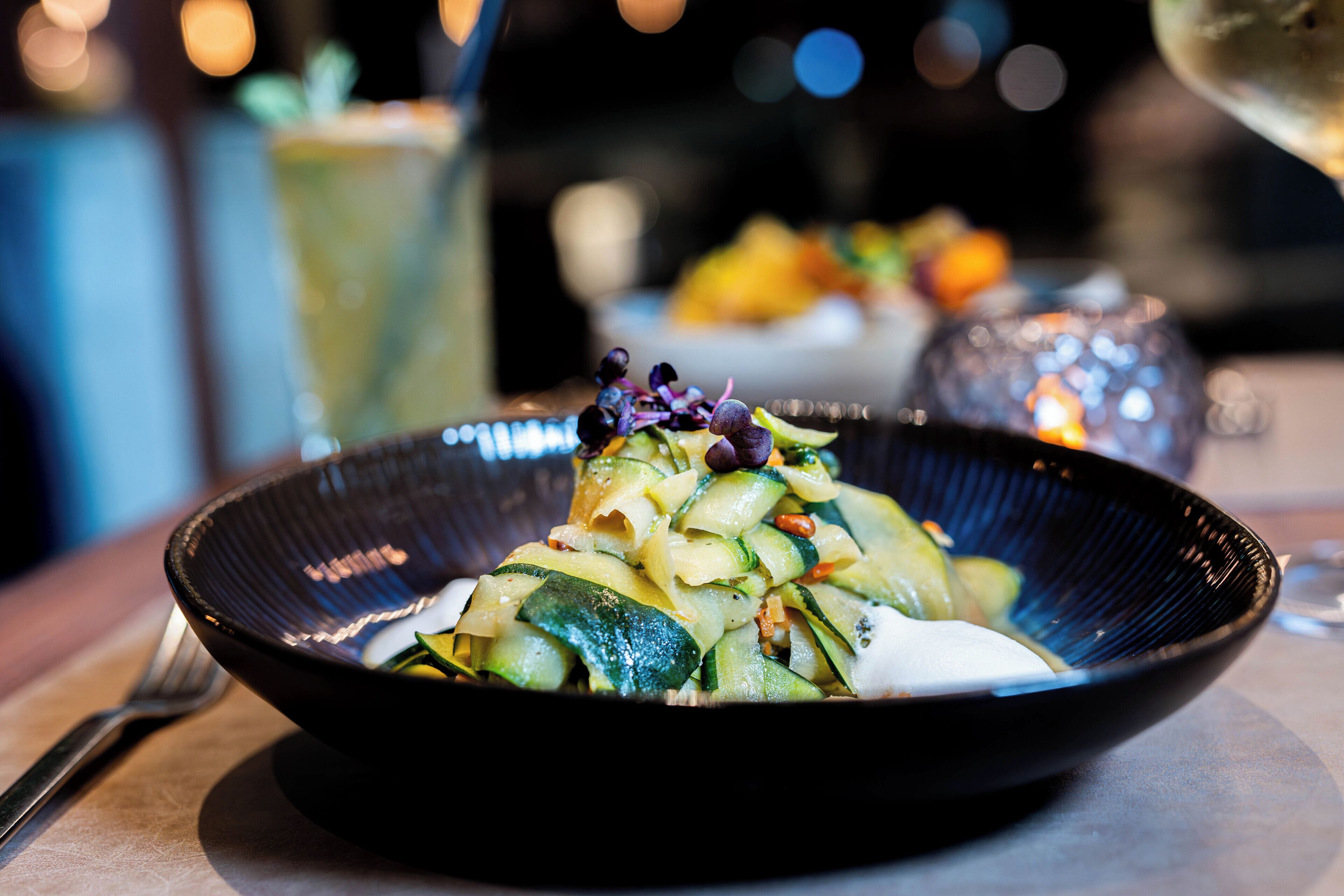 A bowl of pasta with vegetables and a garnish, placed on a table with a glass of wine and a candle in the background.