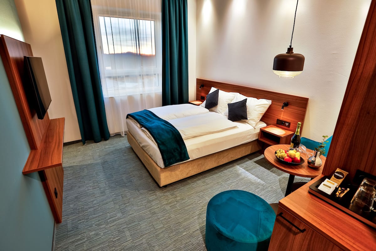 A hotel room with a bed, bedside table, a small round table with a bottle and fruit, a blue ottoman, and a TV mounted on the wall.