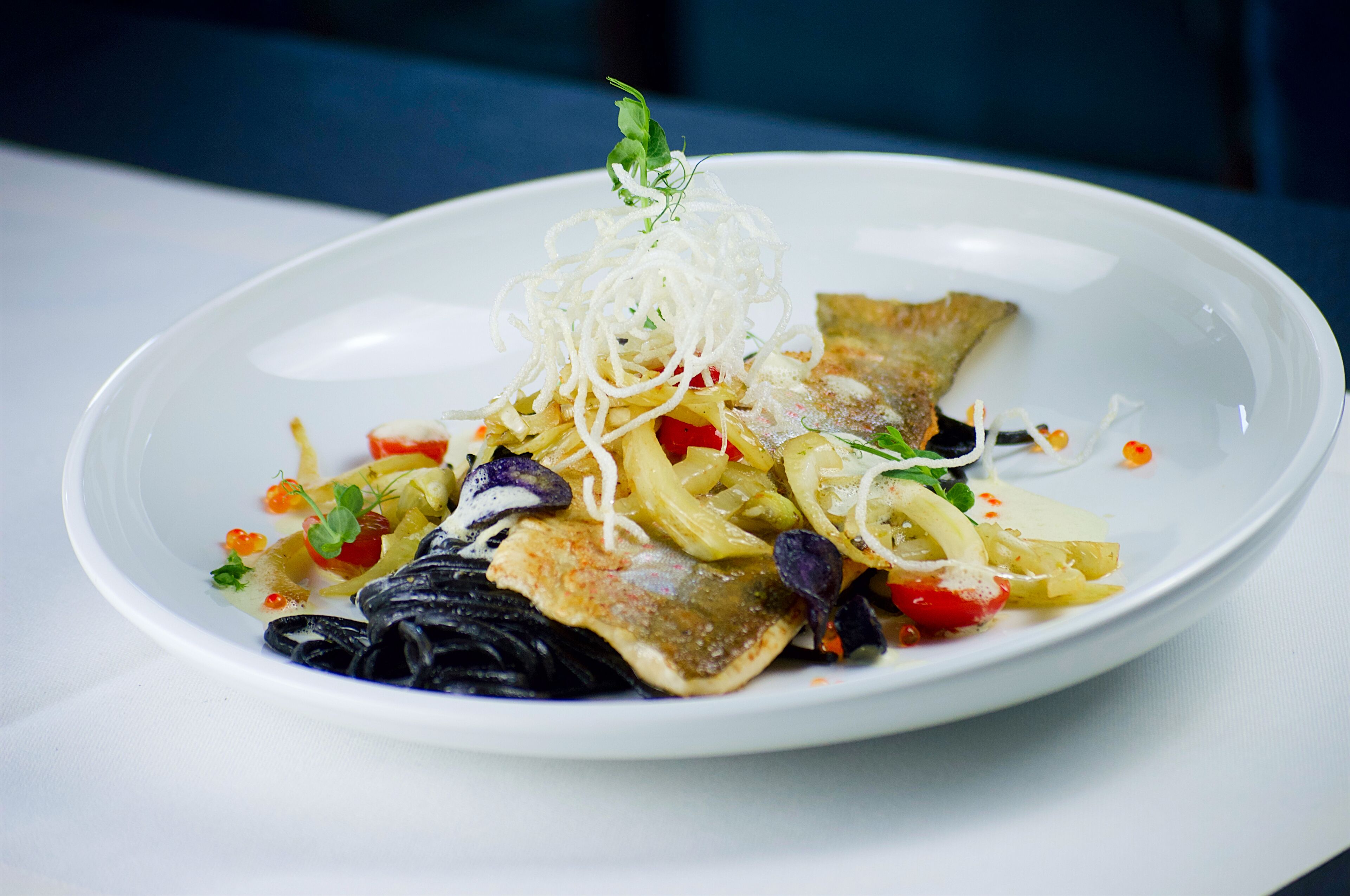Un plato blanco con una variedad de alimentos, incluyendo fideos, verduras y un trozo de pescado, decorado con hierbas.