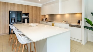 A modern kitchen with a large island, white bar stools, and built-in appliances. - Paradise on the Promontory (BANKSIA BEACH)