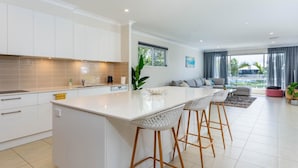 A modern kitchen with white cabinetry, a large island with bar stools, and a dining area with a sofa and coffee table. - Paradise on the Promontory (BANKSIA BEACH)
