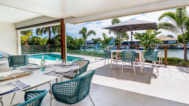 A modern outdoor dining area with a pool view, featuring a table set for two with chairs, a large umbrella, and a glass railing overlooking the pool and greenery. - Paradise on the Promontory (BANKSIA BEACH)