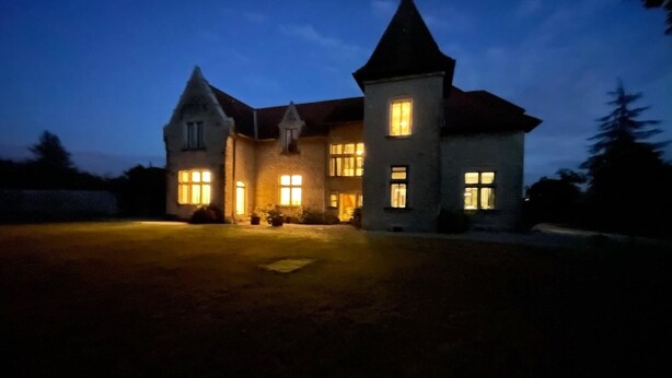A large, two-story house with a prominent tower and multiple lit windows, surrounded by a well-maintained lawn.