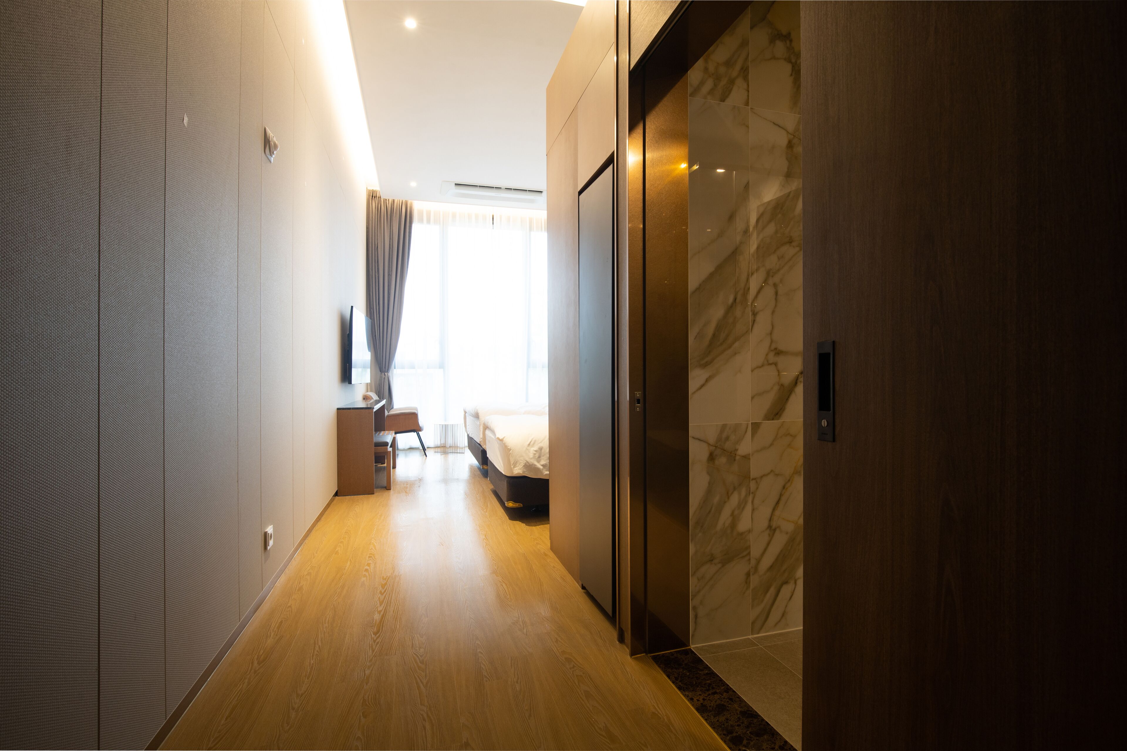 A modern hotel corridor with a wooden floor, a bed, a desk, and a wardrobe.