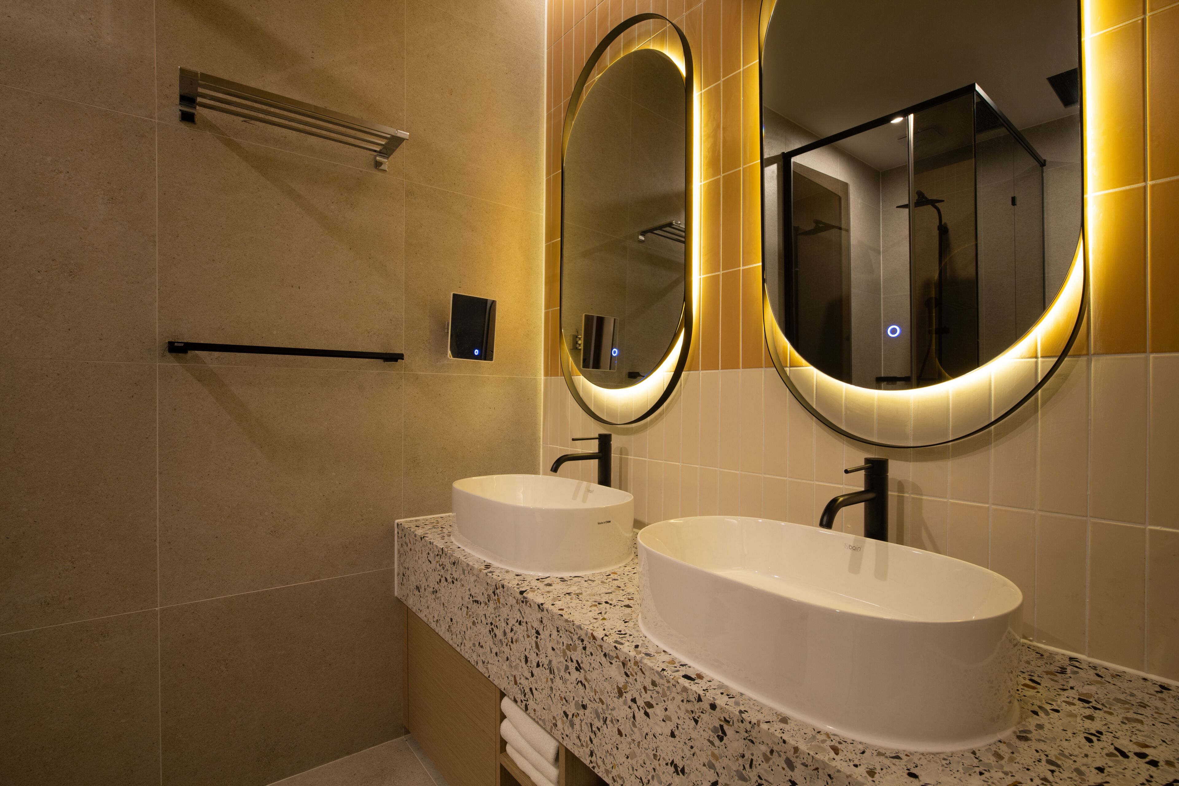 A modern bathroom with two sinks, a towel rack, and two wall-mounted mirrors with illuminated frames.