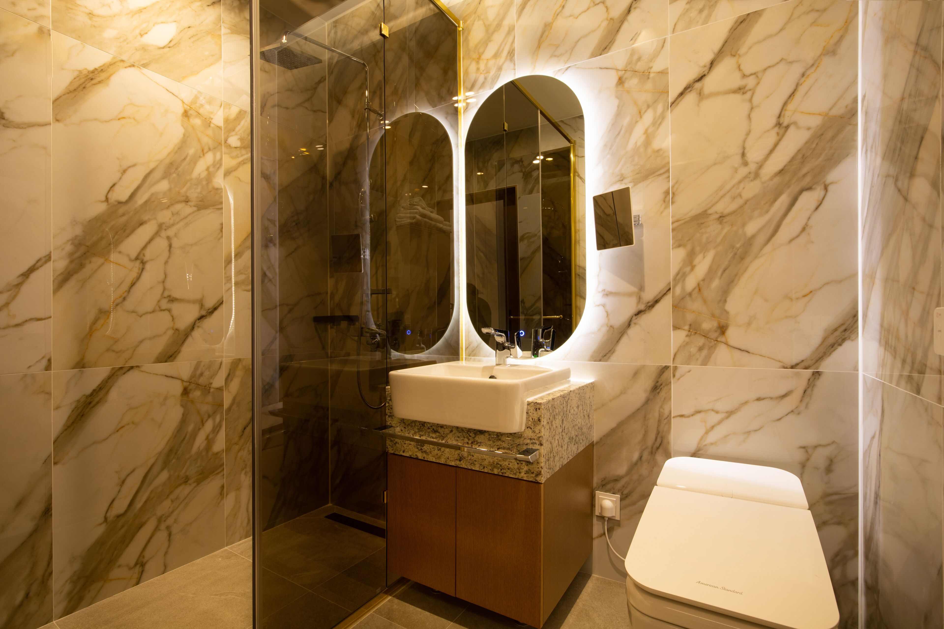 A bathroom with marble walls, a glass shower enclosure, a sink with a mirror, and a toilet.