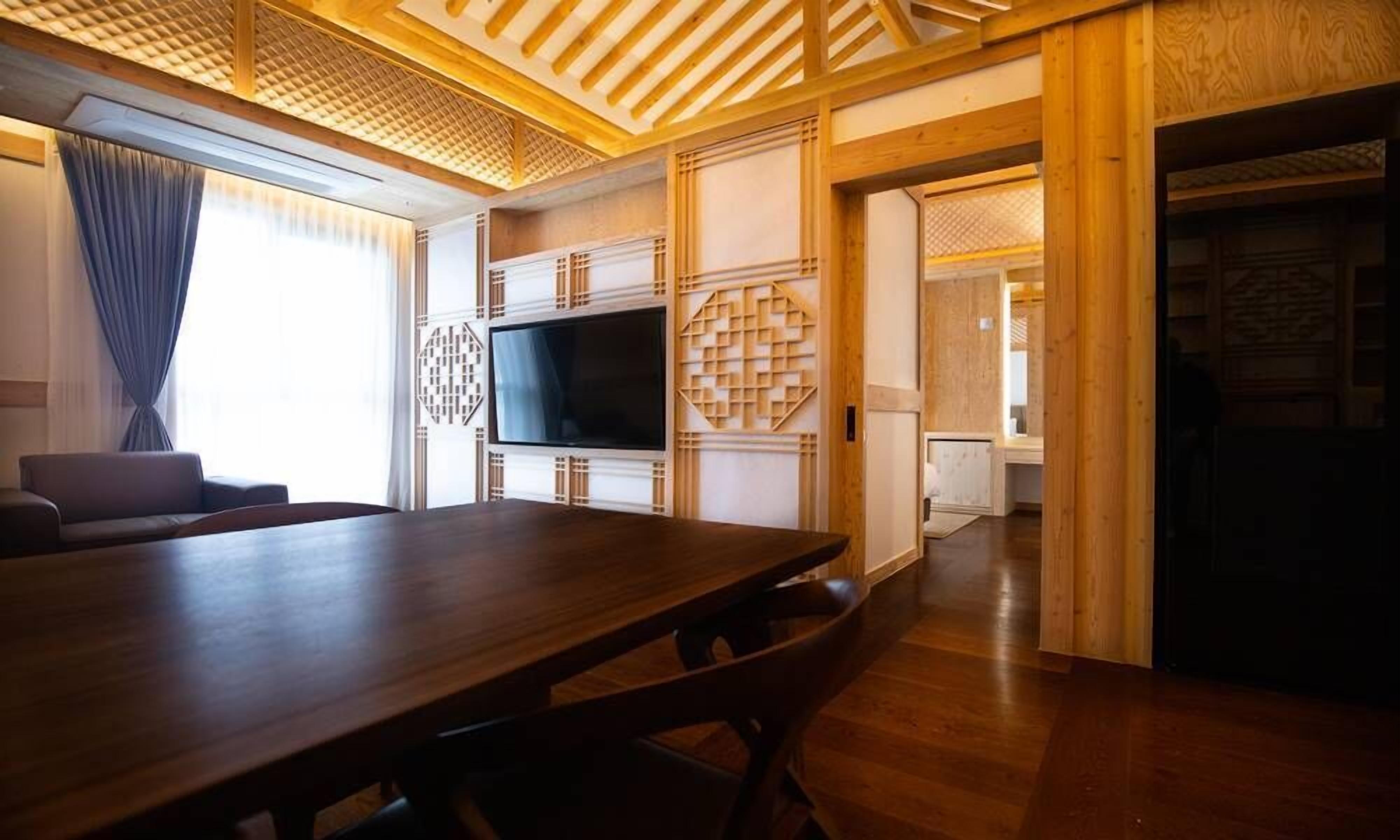 A modern living room with a wooden dining table, a flat-screen TV mounted on a wall, and a sofa.