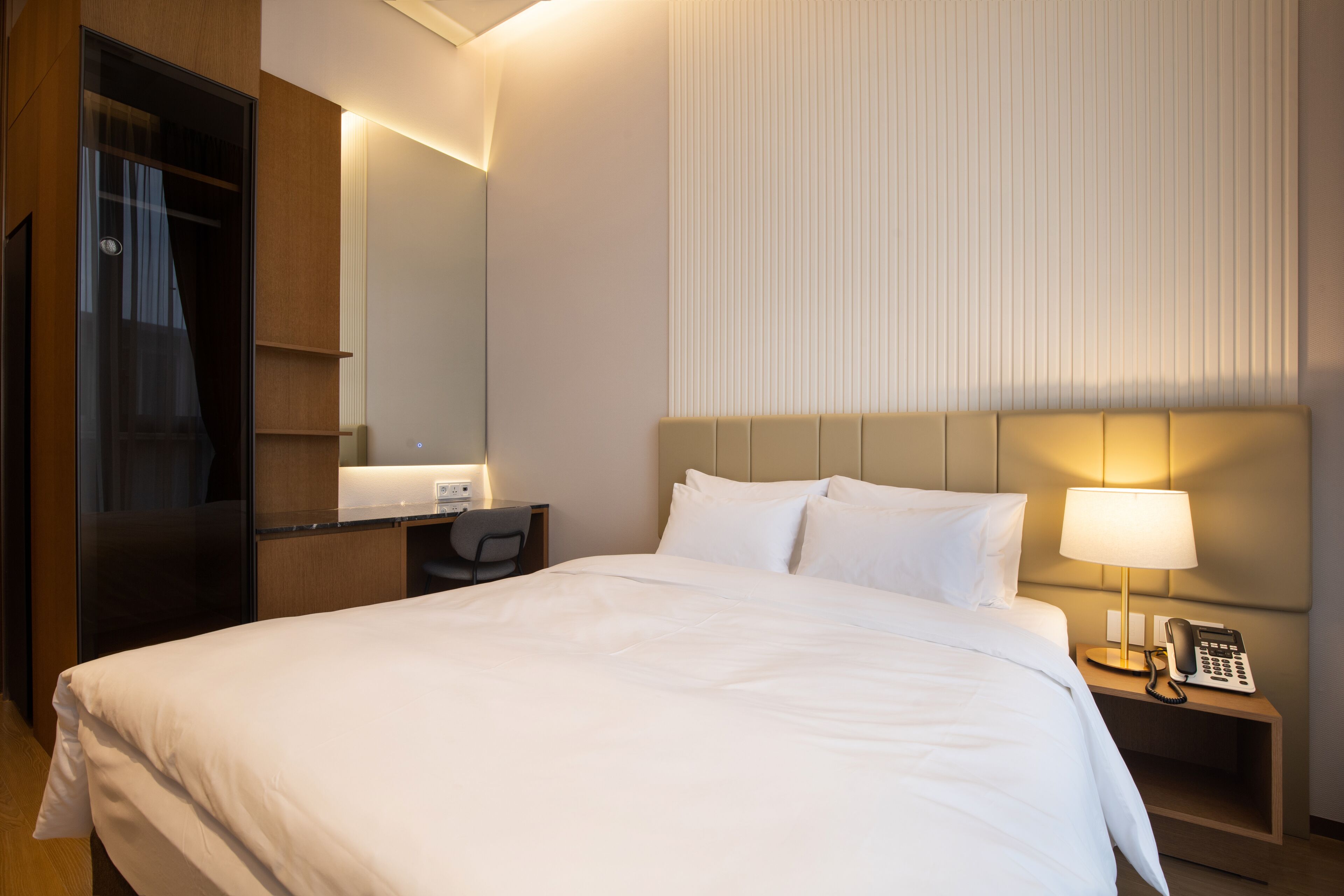 A hotel room with a large bed, a desk with a chair, a bedside lamp, and a telephone.
