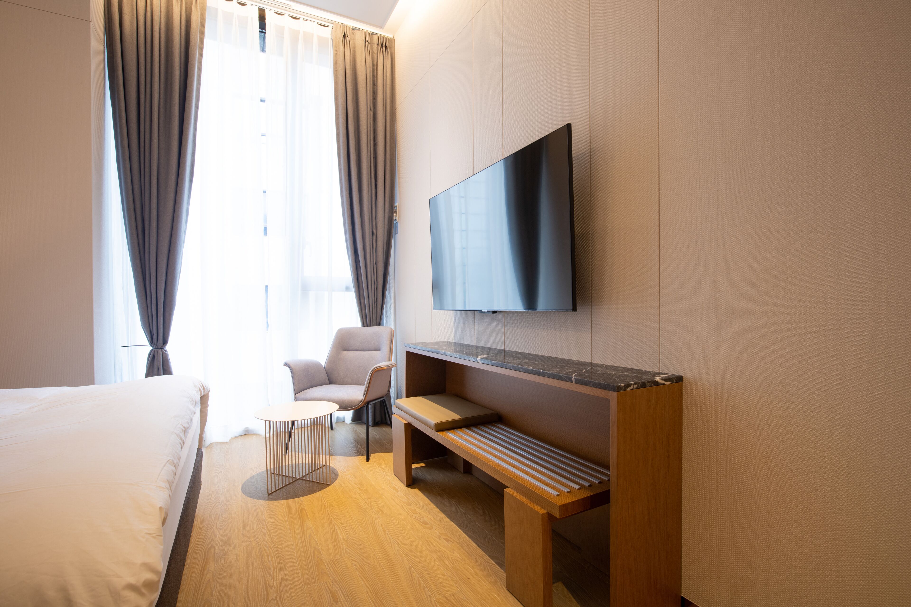 Une chambre d’hôtel moderne dotée d’un grand lit, d’un coin salon avec une petite table, d’un téléviseur à écran plat fixé au mur et d’une grande fenêtre avec des rideaux.