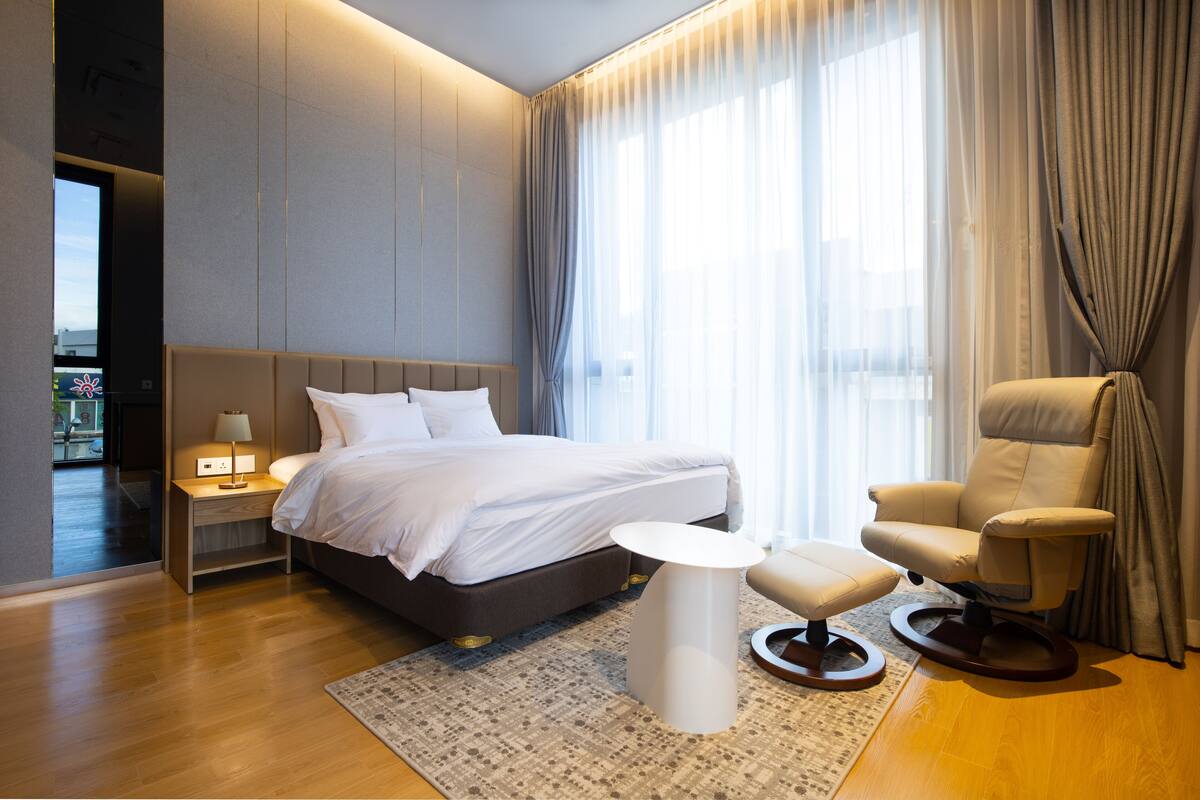 Habitación de hotel moderna con una cama grande, un espacio de descanso con una silla y reposapiés, una mesita auxiliar y vistas al exterior a través de cortinas translúcidas.