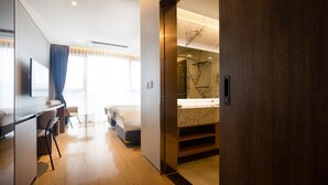 Een moderne hotelkamer met een houten vloer, een bureau met stoelen, een bed, een badkamer met een marmeren wastafel en een grote spiegel.