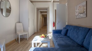 Ein modernes Wohnzimmer mit einem blauen Sofa, einem weißen Sessel und einem kleinen Tisch mit Obst. Ein Flur führt zu einem weiteren Raum.