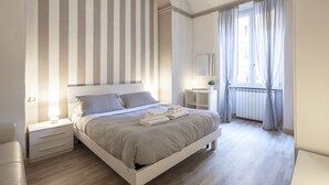 Ein Schlafzimmer mit einem großen Bett, einem Nachttisch, einem Radiator, einem Fenster mit Vorhängen und einem Spiegel an der Wand.