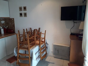 Uma área de refeições com mesa redonda e várias cadeiras, uma cozinha com uma torradeira vermelha e uma televisão na parede.