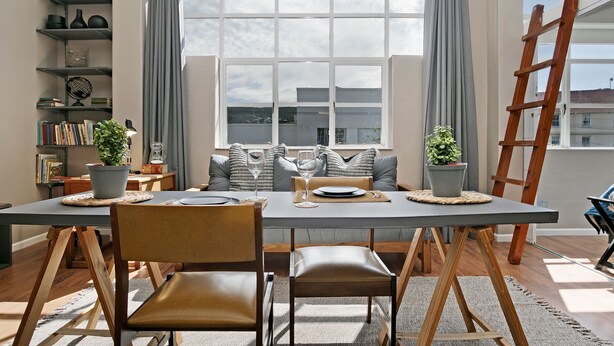 A dining area with a table set for two, a bookshelf, and a window with curtains. - Studio on Hope (Cape Town)