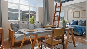 A modern living room with a dining area, large windows, a loft bed, and a ladder. - Studio on Hope (Cape Town)