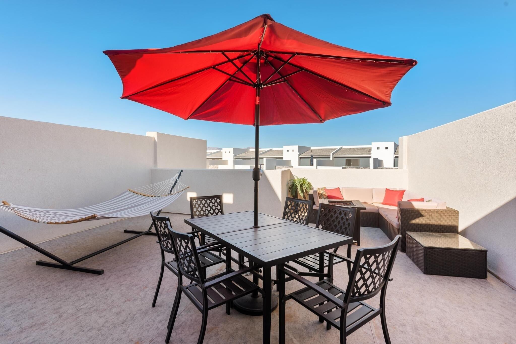 A rooftop terrace with a table and chairs, a red umbrella, a hammock, and a sofa with cushions.