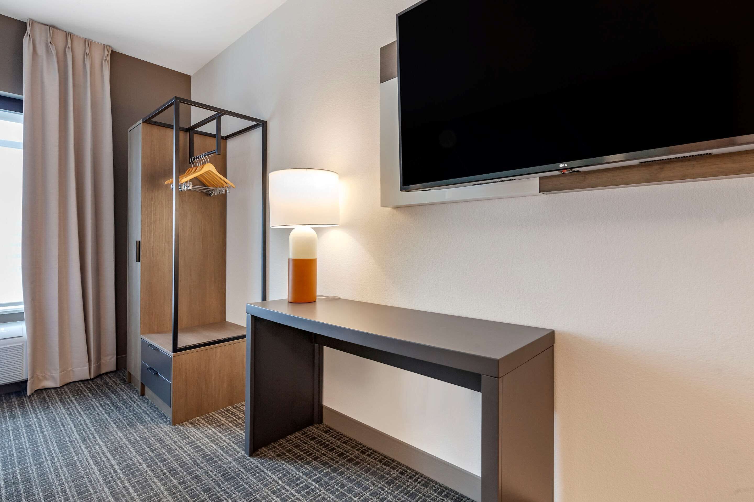 A hotel room with a large flat-screen TV mounted on the wall, a wooden wardrobe with a hanging rack, a desk with a lamp, and a patterned carpet.
