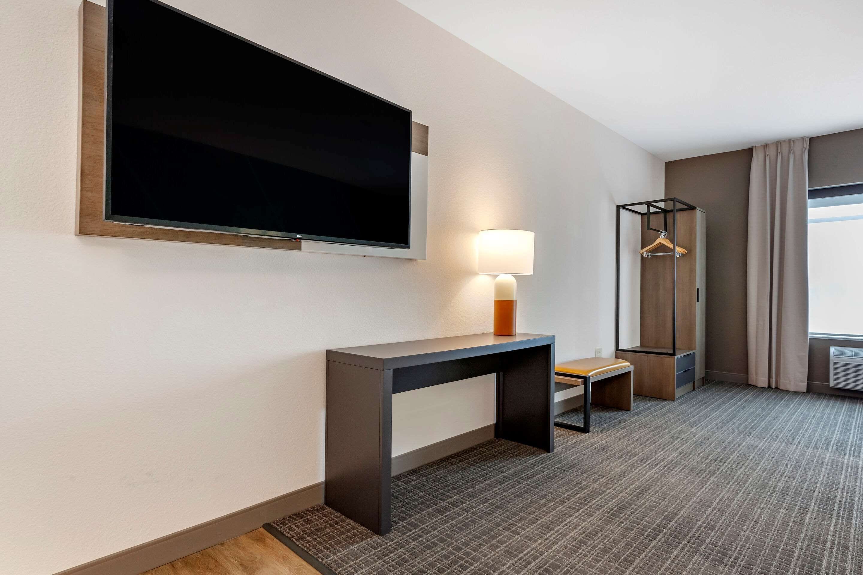 Habitación de hotel con una televisión de pantalla plana grande montada en la pared, una mesita de noche con lámpara, un pequeño banco y un perchero.