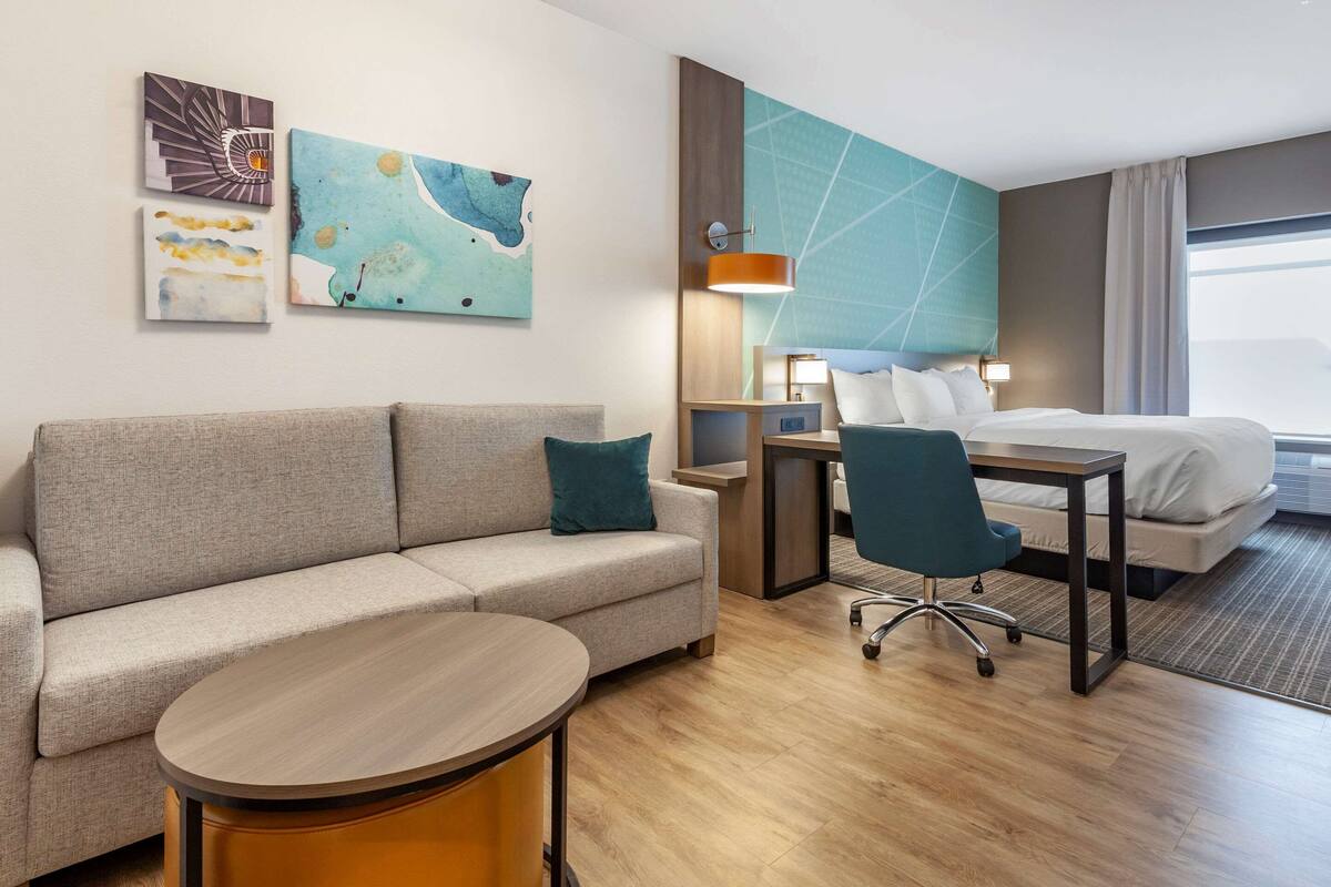 A hotel room with a sofa, a round table, a desk with a chair, a bed, and artwork on the walls.
