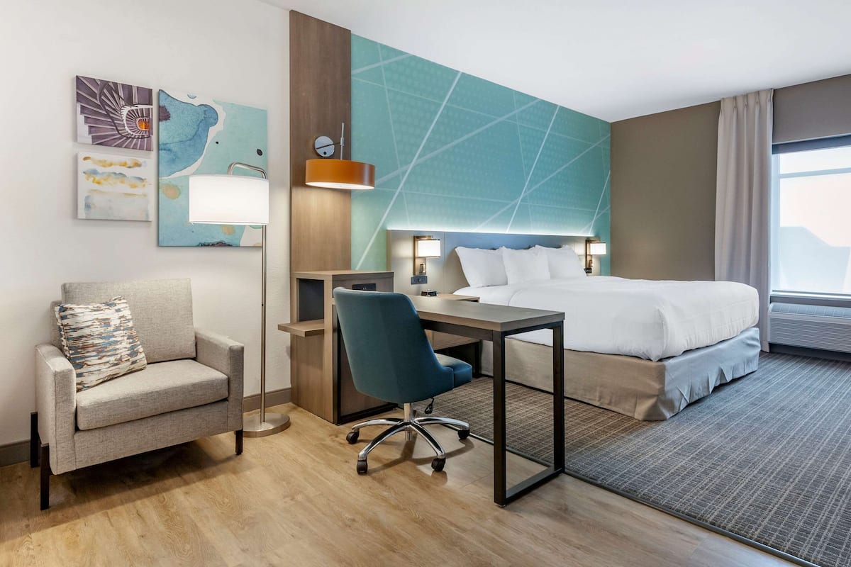 A hotel room with a bed, desk, chair, and artwork on the wall.