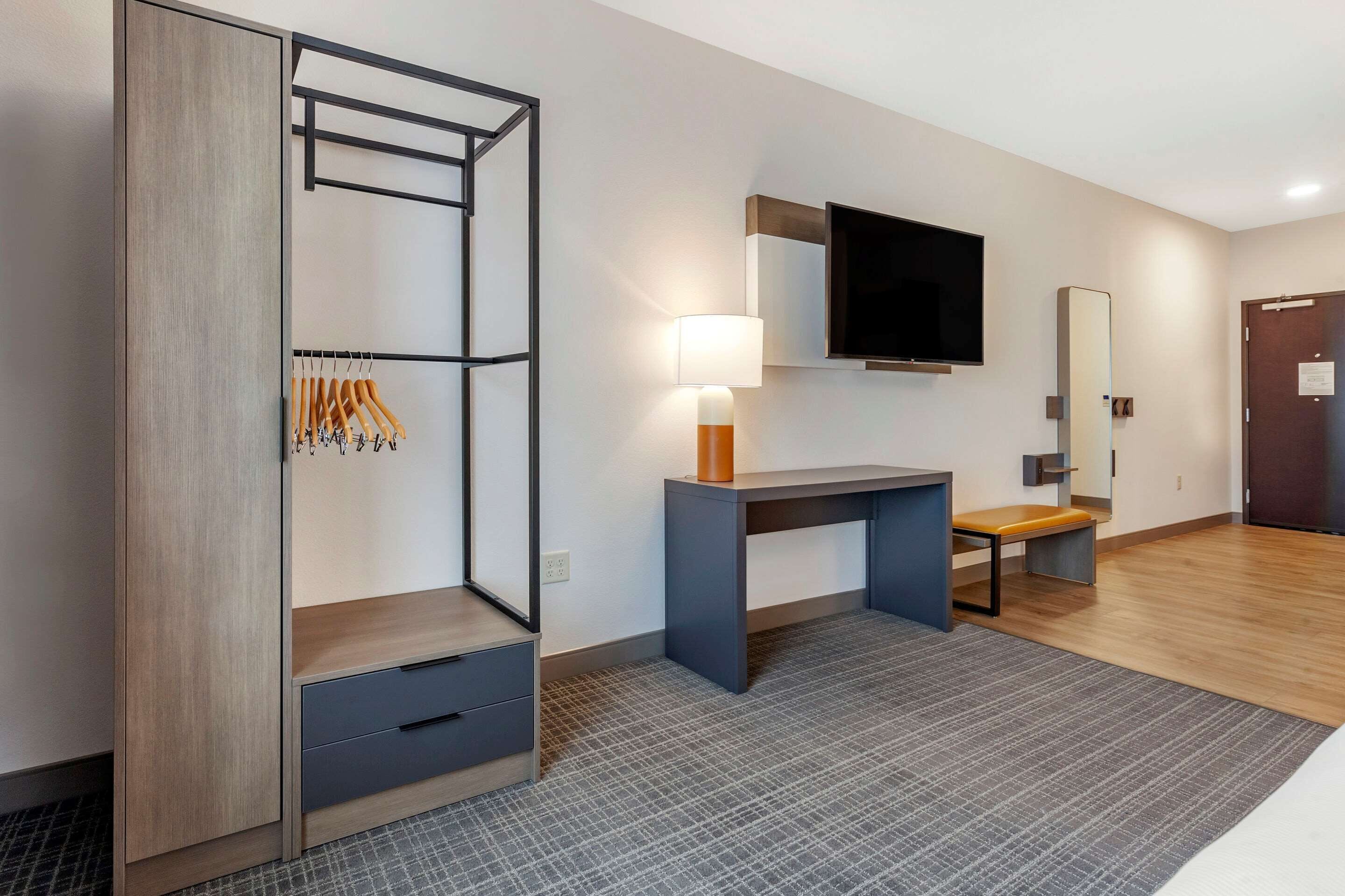 Une chambre d’hôtel moderne équipée d’un placard, d’un bureau avec une lampe, d’un téléviseur à écran plat et d’un banc.