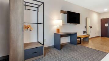 A modern hotel room with a wardrobe, a desk with a lamp, a flat-screen TV, and a bench.
