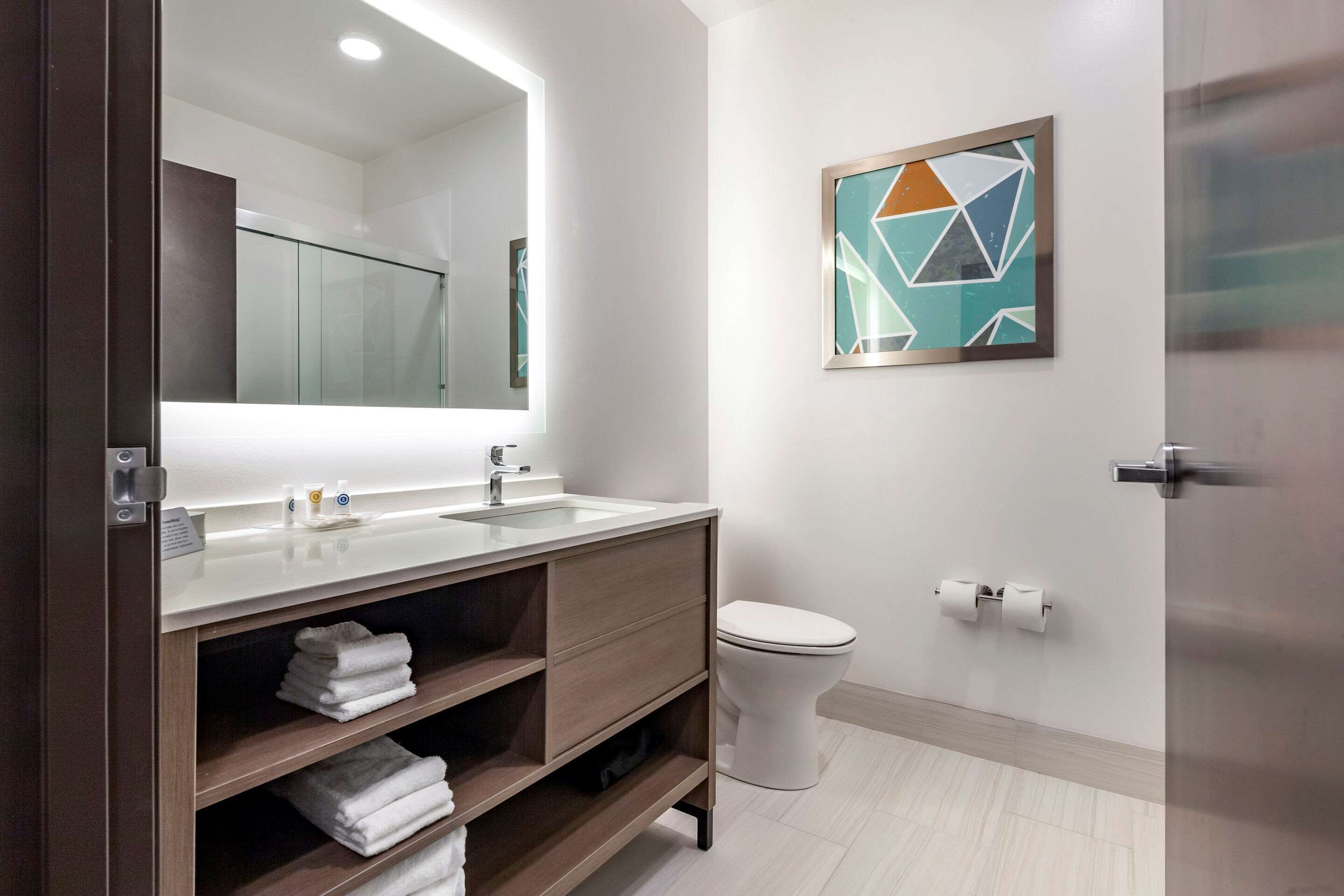 Un baño moderno con tocador de madera, un espejo grande, un inodoro y una obra de arte geométrica en la pared.