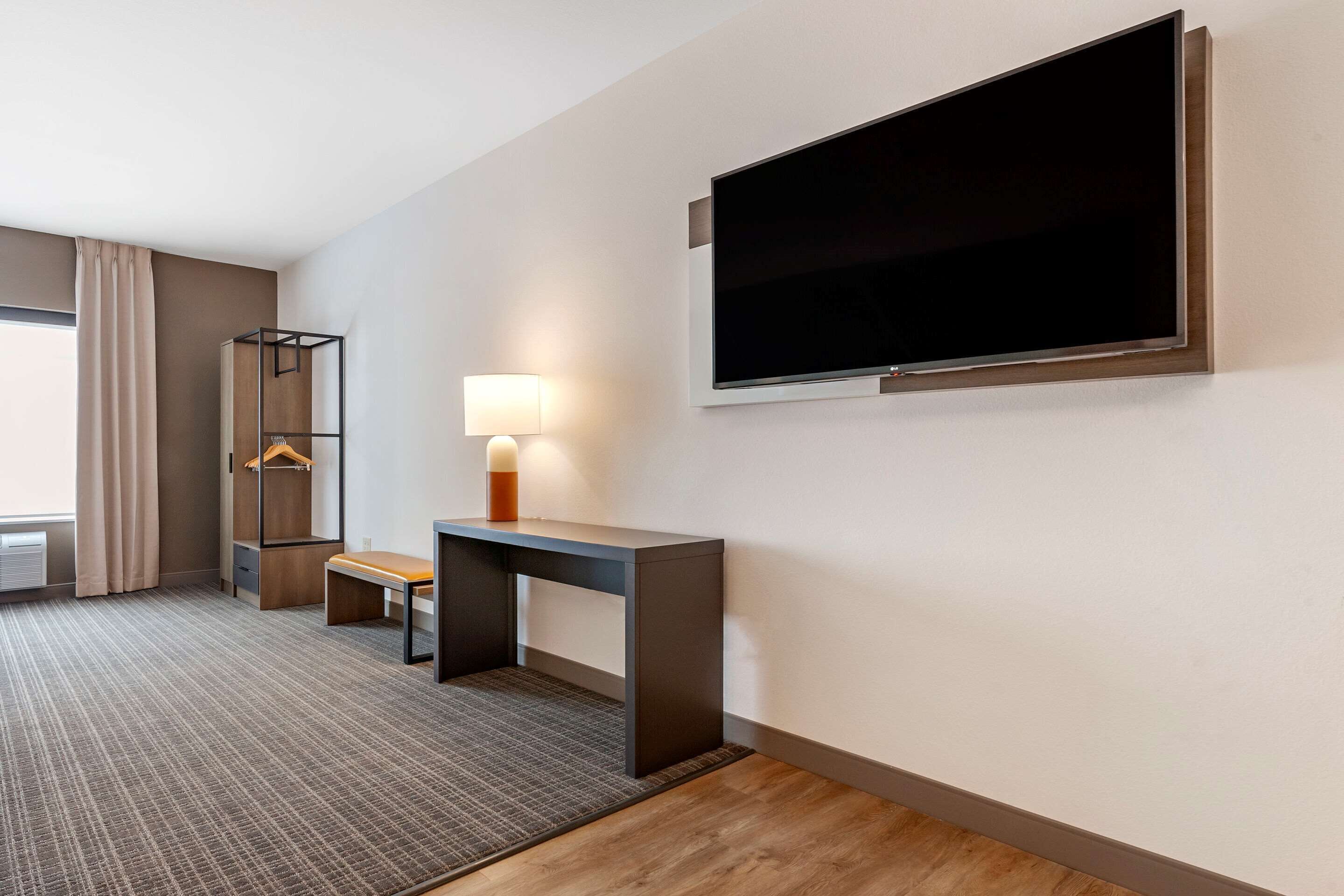 A hotel room with a bed, a desk with a lamp, a TV mounted on the wall, and a window with curtains.