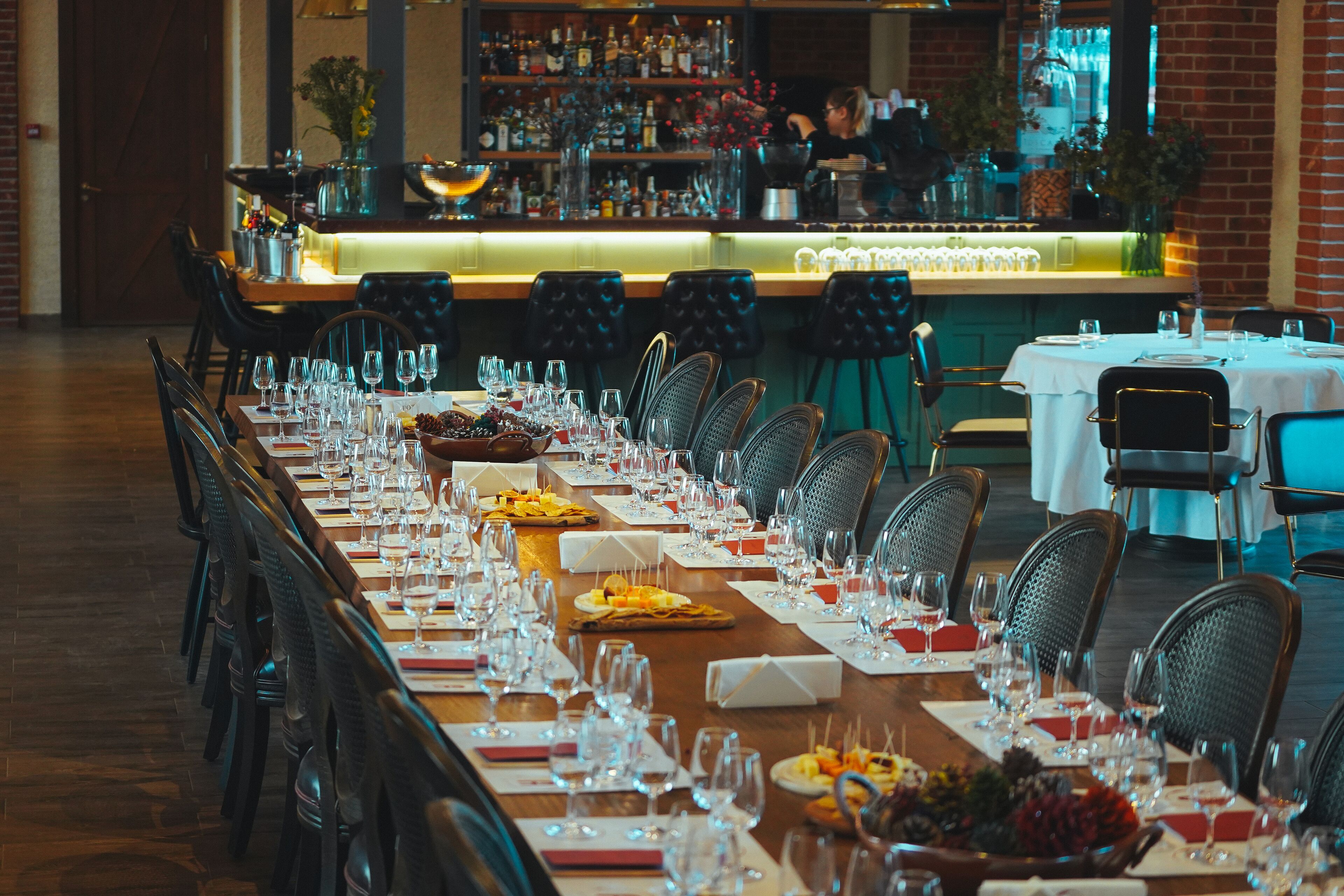 Una larga mesa de comedor dispuesta con múltiples sillas, una zona de bar al fondo y una pared de ladrillo.
