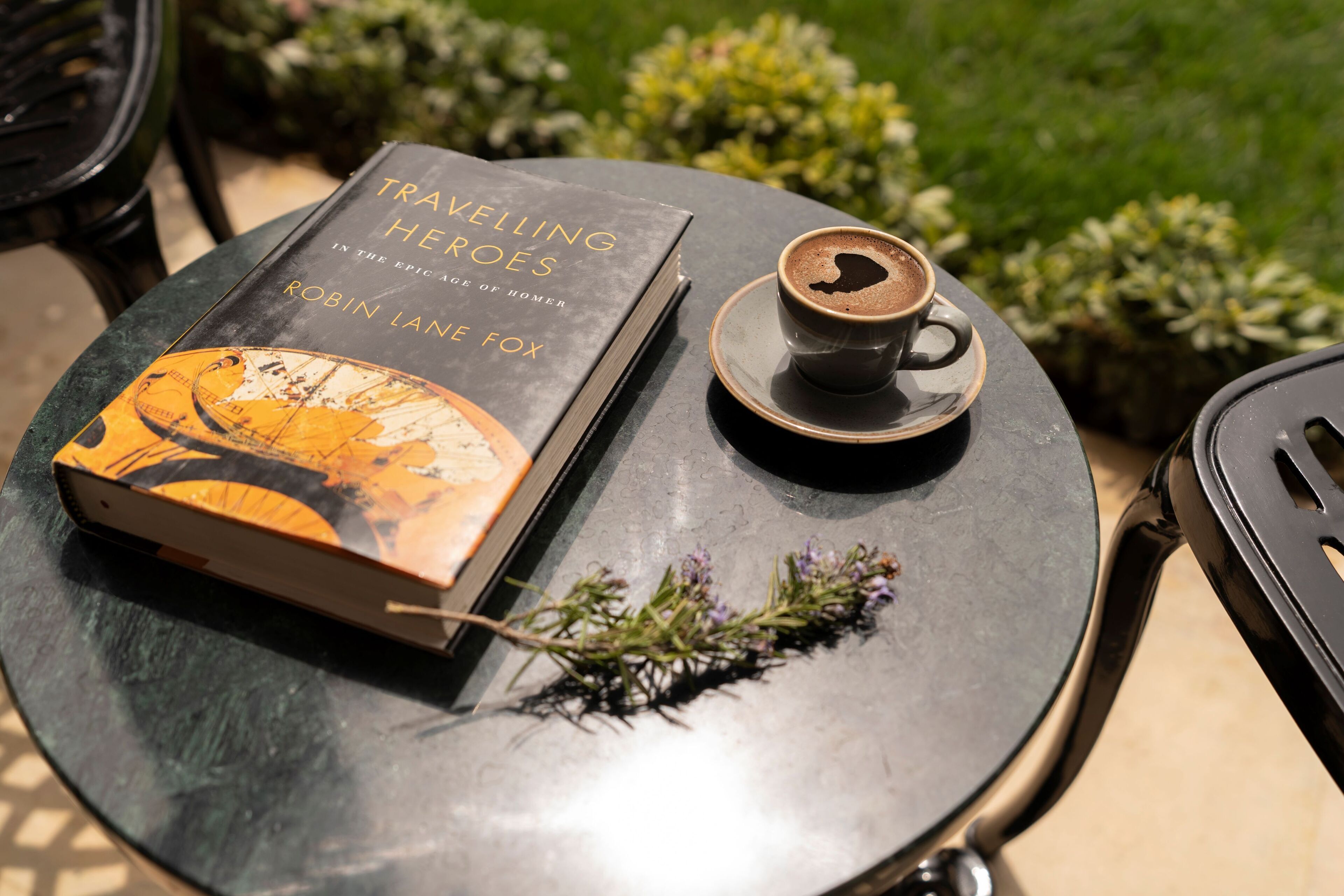 A book titled 'Travelling Heroes' by Robin Lane Fox on a round table with a cup of coffee and a sprig of lavender.