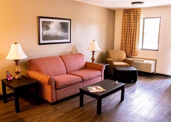 A hotel room with a sofa, two lamps, a coffee table, a chair, a radiator, and a window with curtains.