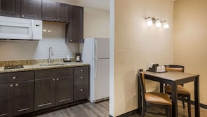 A modern kitchen with dark cabinets, a white microwave, and a refrigerator. There is a dining area with a table and chairs, and a wall-mounted light fixture. - Econo Lodge Vidalia (Vidalia)