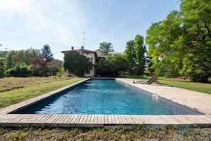 A rectangular swimming pool with a wooden deck, surrounded by greenery and a building in the background.