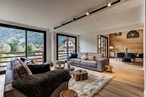 A modern living room with a sofa, a dining area with a table and chairs, and a large window offering a view of a mountain landscape. - Luxe La Cordee 221 penthouse - Chamonix All Year (Chamonix-Mont-Blanc)