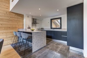 A modern kitchen with a central island, bar stools, and a window overlooking a wooden structure. - Luxe La Cordee 221 penthouse - Chamonix All Year (Chamonix-Mont-Blanc)