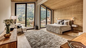 A bedroom with a large bed, a chair, a desk, and a view of the mountains through large windows. - La Cordee 221 Penthouse -  sleeps 8 guests with pool, sauna, gym, parking (Chamonix-Mont-Blanc)