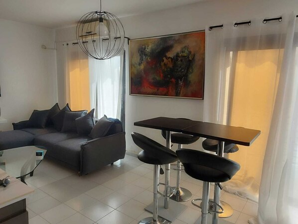 A modern living room with a sectional sofa, a dining table with bar stools, a large abstract painting, and a globe chandelier.