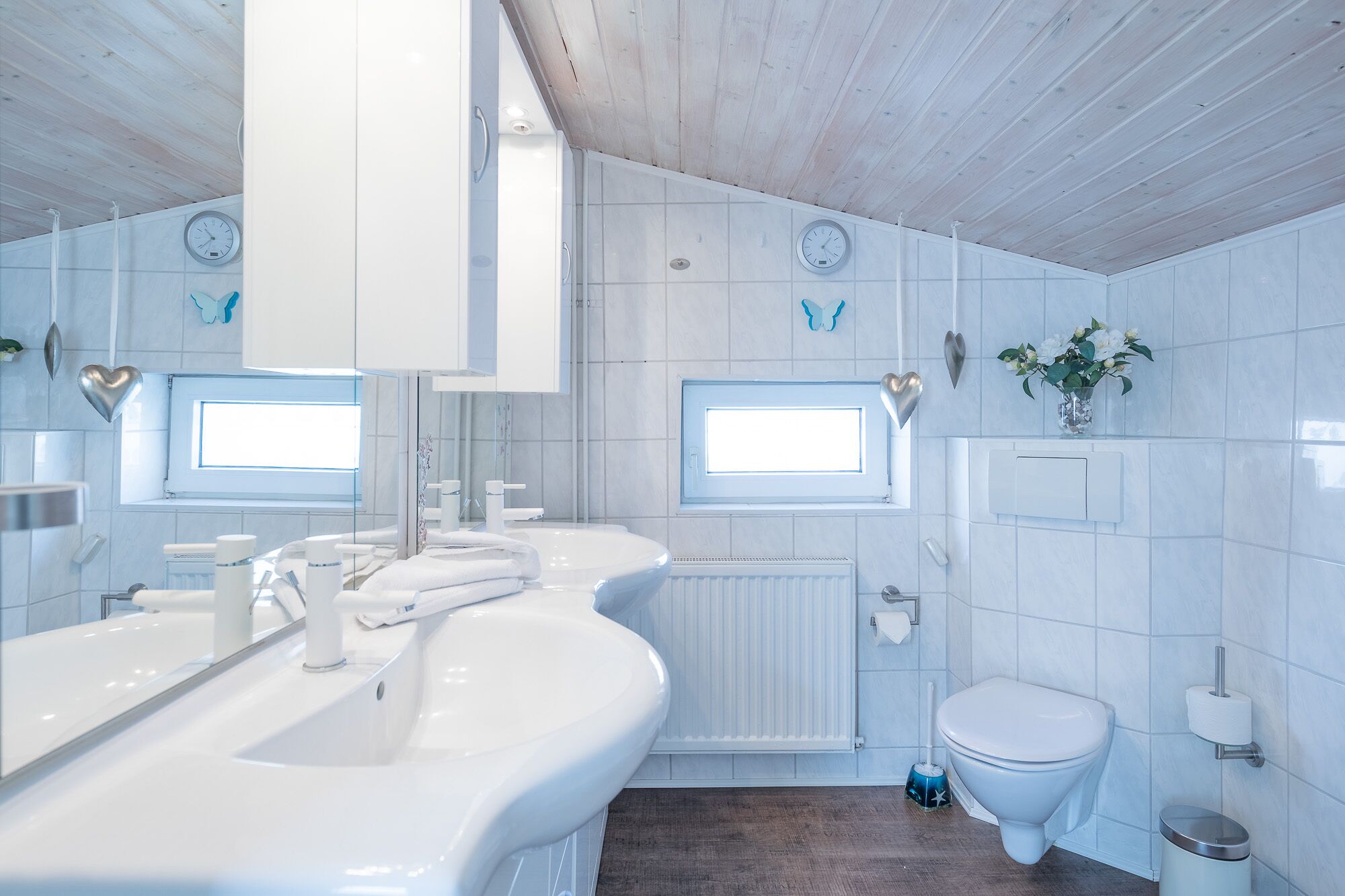 Ein modernes Badezimmer mit einem weißen Waschbecken, einer Toilette und einem Waschtisch. Zwei Wanduhren, ein Fenster und eine kleine Pflanze sind an der Wand angebracht.