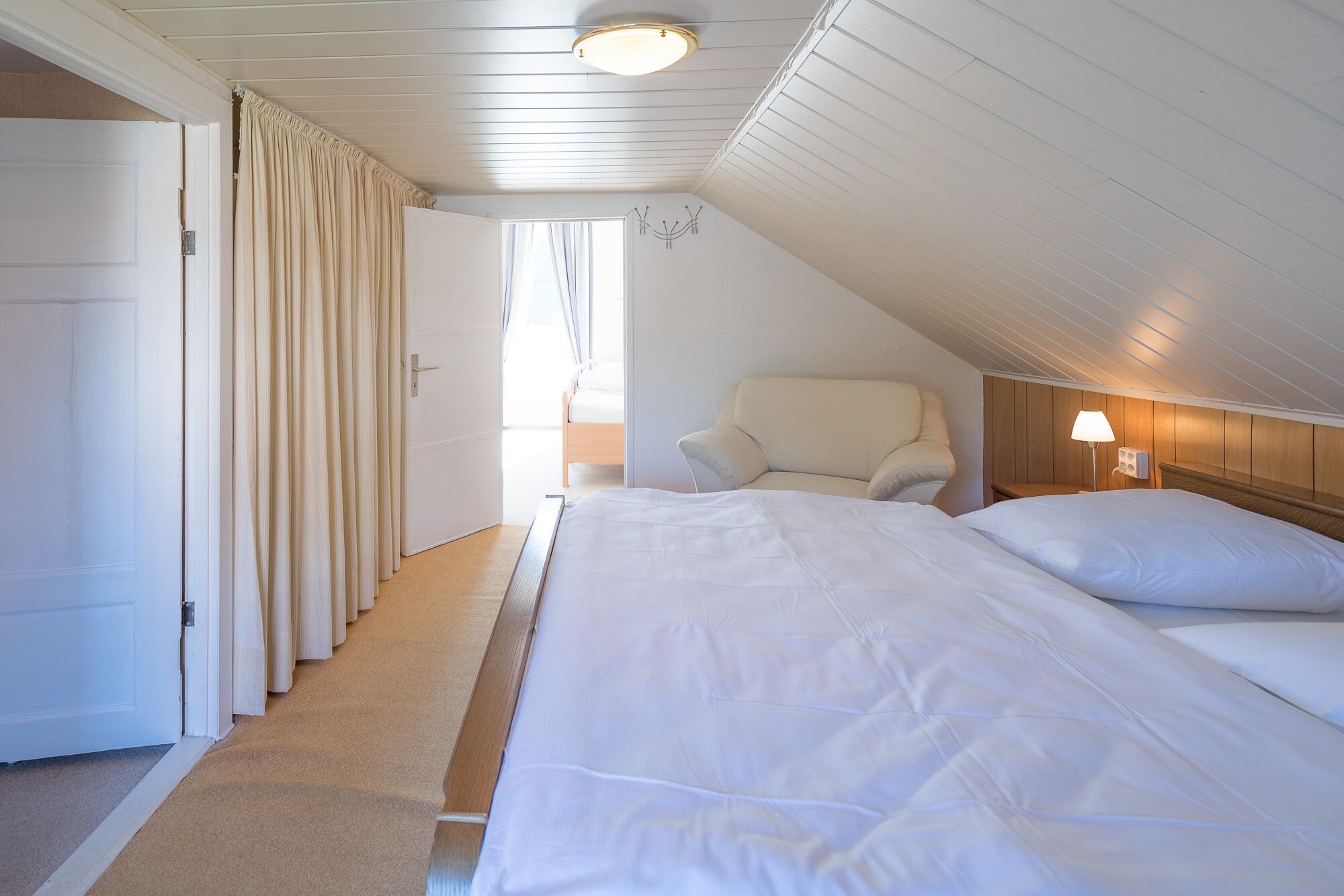 A bedroom with a large bed, a chair, a bedside table with a lamp, and a door leading to another room.