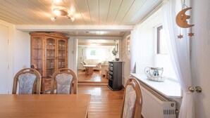 Ein Essbereich mit Holztisch und Stühlen, einem Schrank mit Gläsern und Blick in ein Wohnzimmer mit Sofa und Tisch.