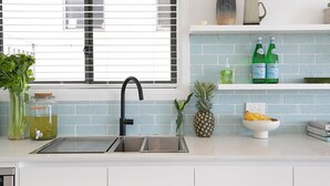 Een moderne keuken met een roestvrijstalen spoelbak, een raam met zonwering, een blauwe tegelwand en diverse keukengerei op het aanrecht.