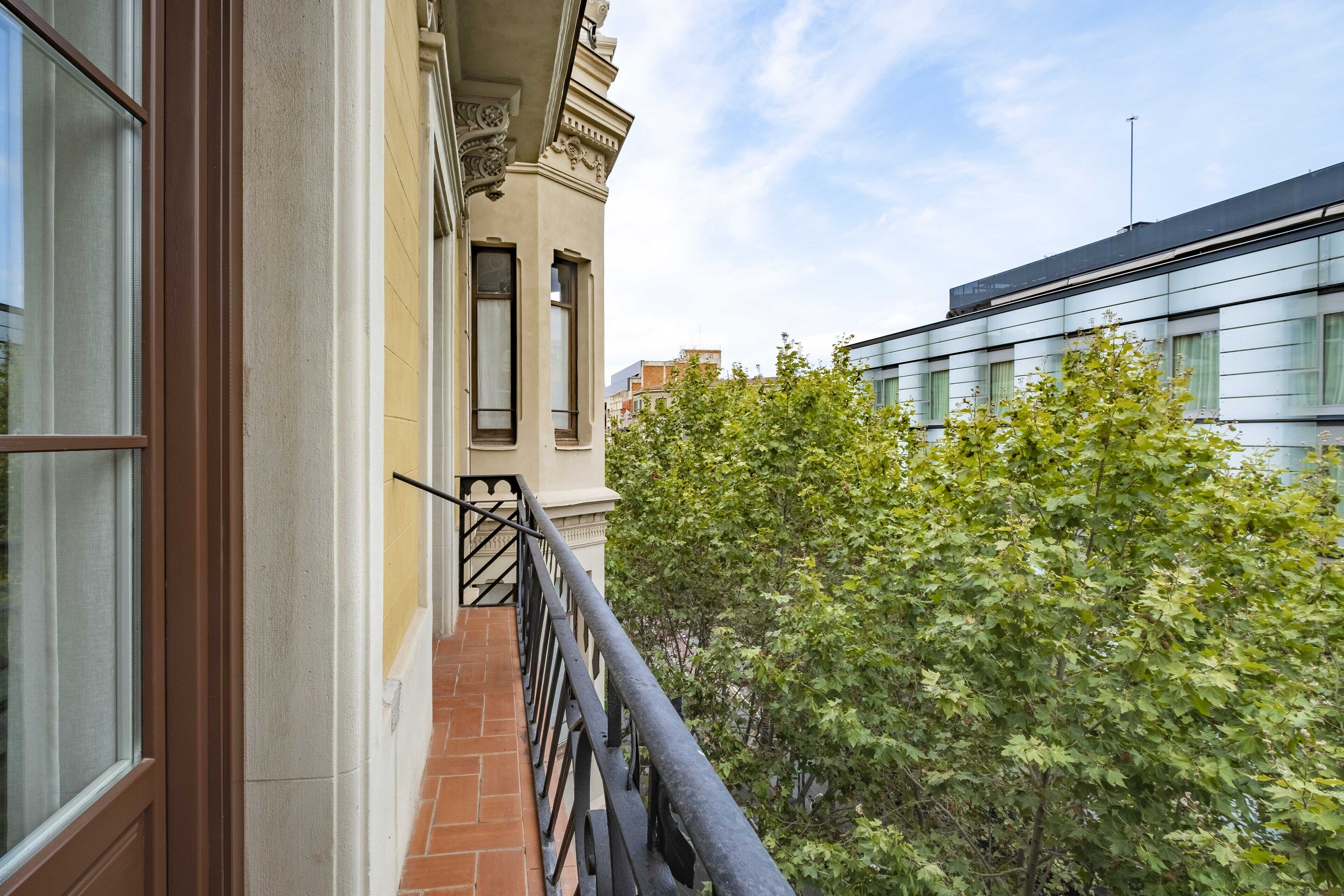 A balcony with a wrought iron railing, a brick floor, and a view of a tree and neighboring buildings.
