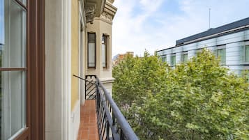 A balcony with a wrought iron railing, a brick floor, and a view of a tree and neighboring buildings.
