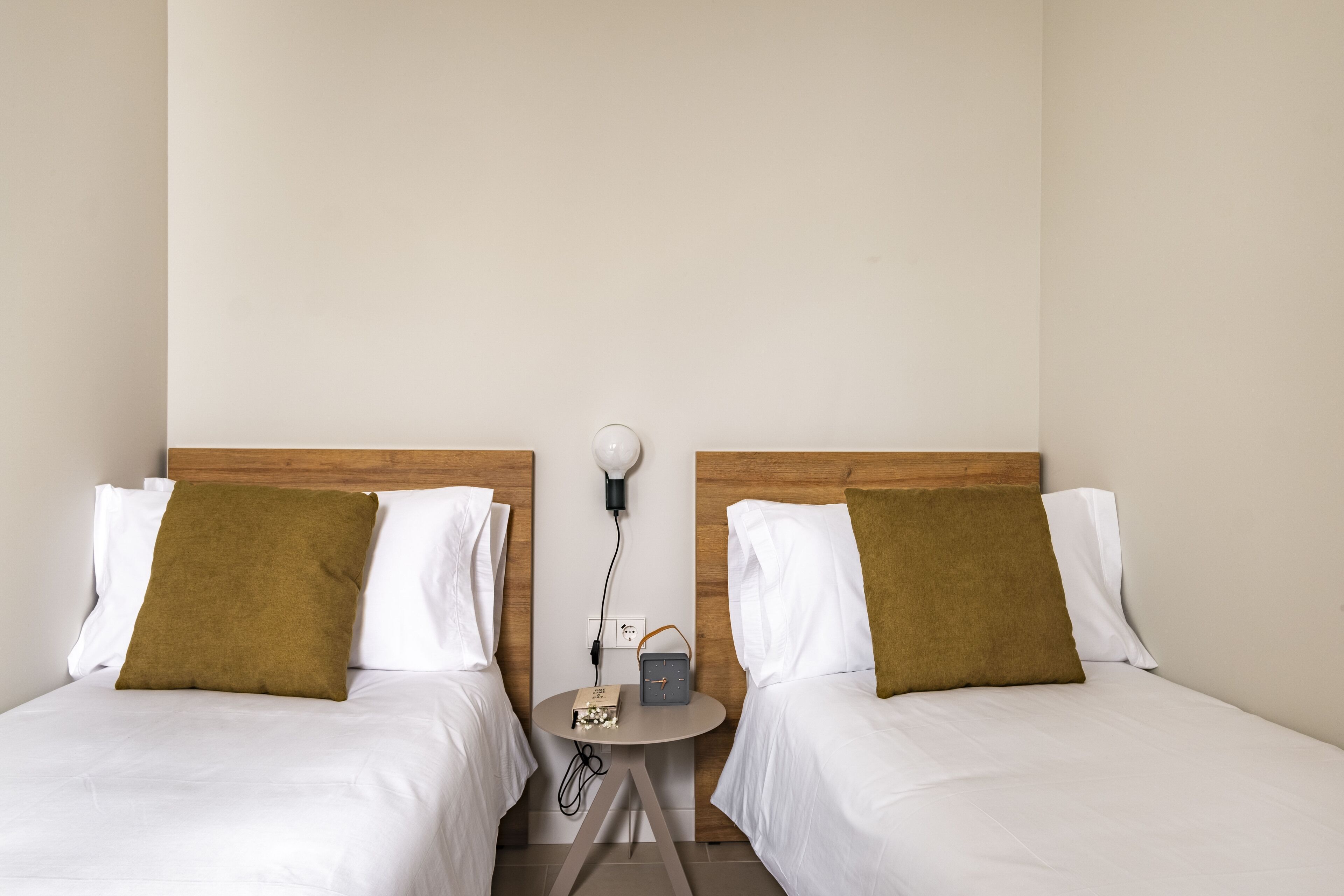 Une chambre d’hôtel avec deux lits, chacun étant équipé de draps blancs et d’un oreiller marron. Une petite table ronde se trouve entre les lits, sur laquelle sont posés une horloge et une lampe.