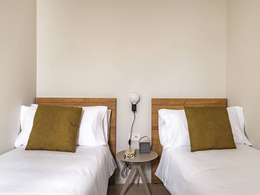 Une chambre d’hôtel avec deux lits, chacun étant équipé de draps blancs et d’un oreiller marron. Une petite table ronde se trouve entre les lits, sur laquelle sont posés une horloge et une lampe.
